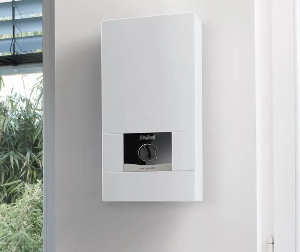Weißer Elektro-Durchlauferhitzer von Vaillant, wandmontiert mit einstellbarem Regler und einem klaren Design, platziert an einer hellen Wand neben einer Fensterscheibe.